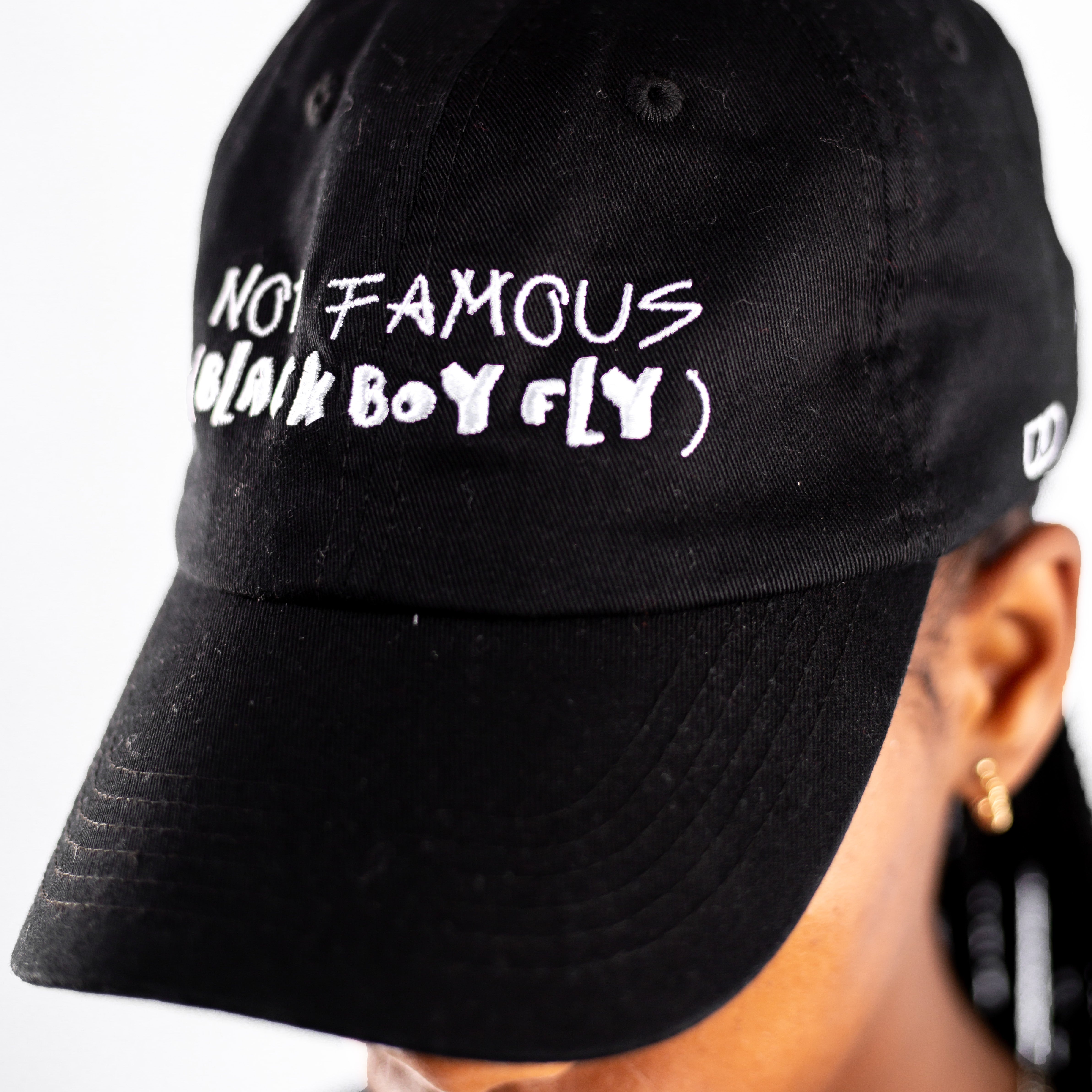 Black baseball cap with white embroidered text reading “NOT FAMOUS BLACK BOY FLY” on the front, shown worn on a person.