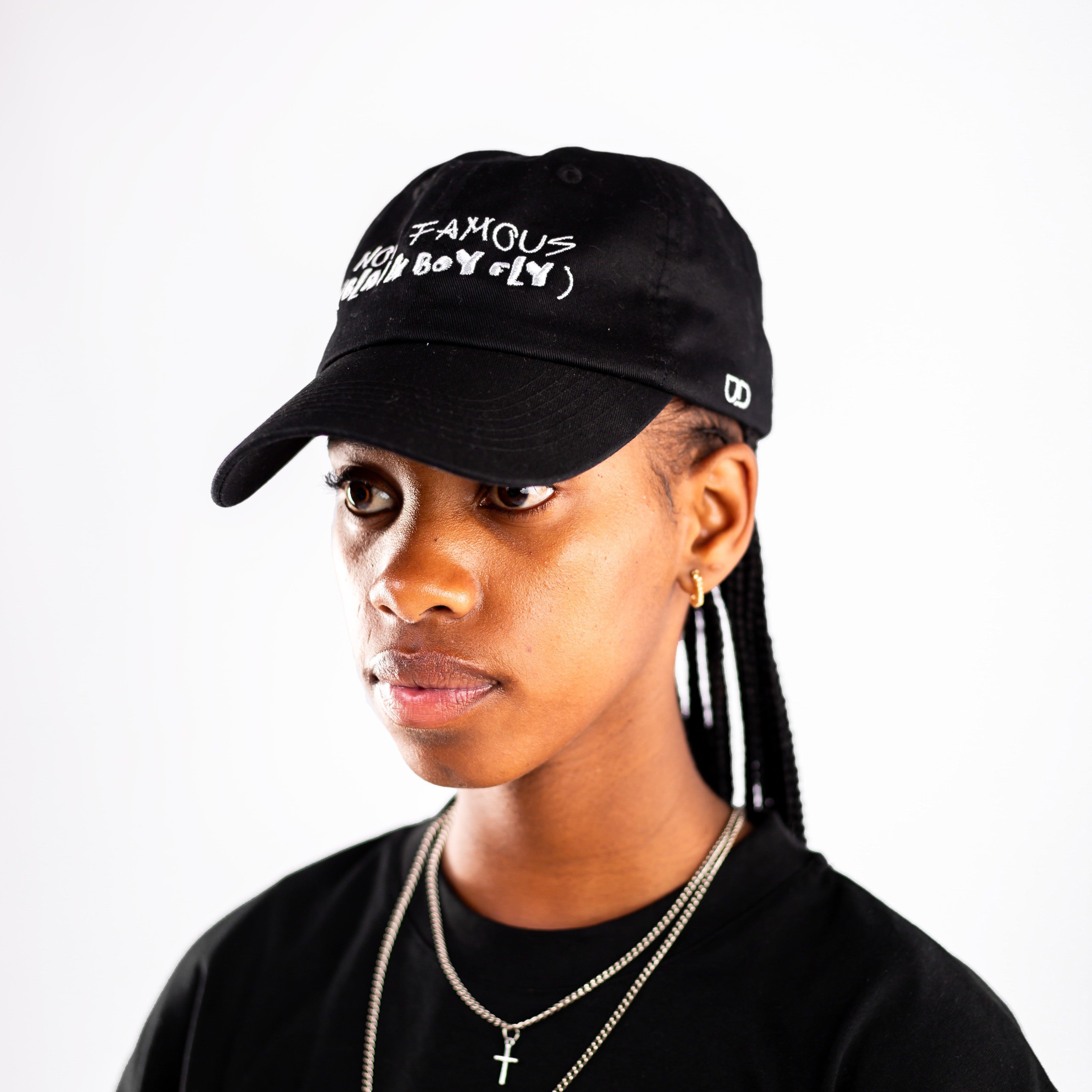 Black cap with white embroidered text reading “NOT FAMOUS (BLACK BOY FLY)”, worn by a person with necklaces and a black shirt