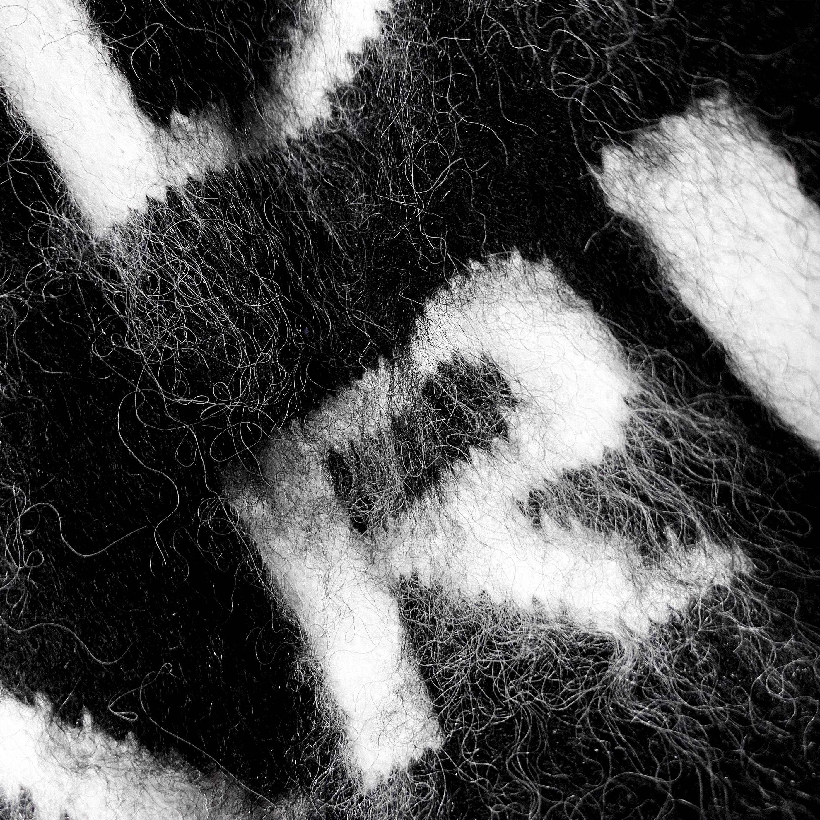 Close-up view of fuzzy black and white knitwear fabric, showing intricate texture and partial white letter or symbol patterns against a black background.