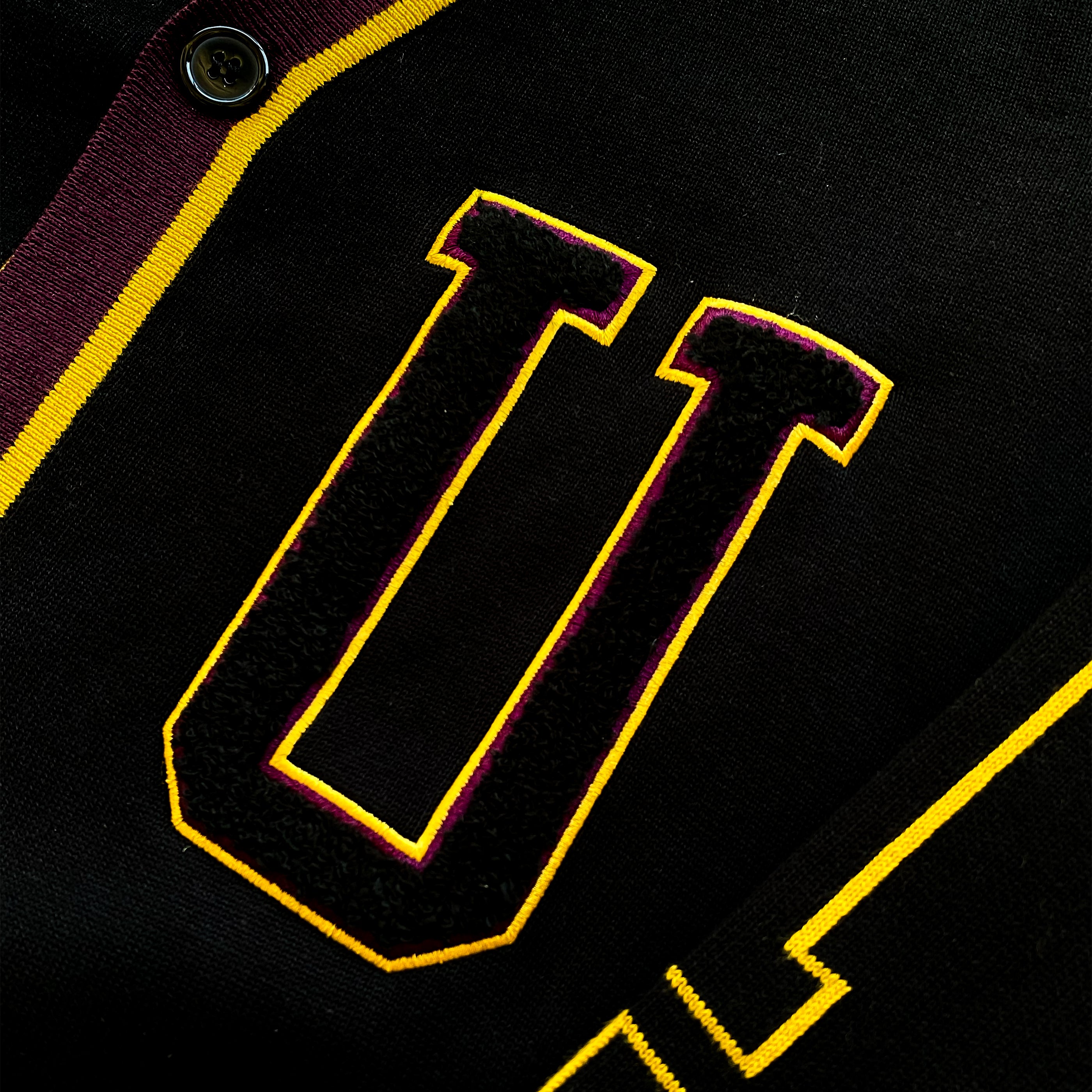 Close-up of a black varsity jacket featuring a large yellow and maroon stitched letter "U" on the chest, with button and collar details visible.