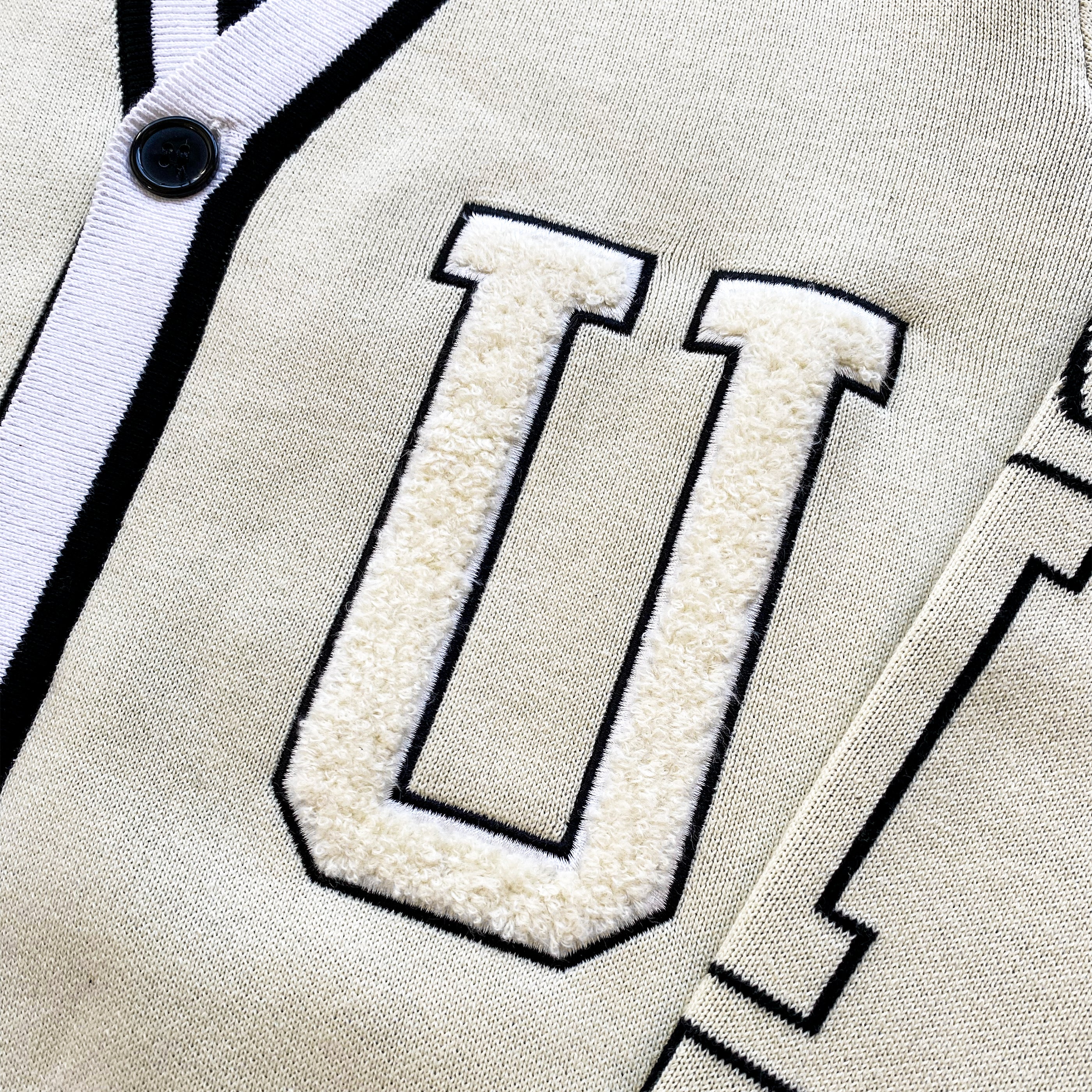 Close-up of the letter “U” applique on the cream white varsity-style cardigan with black trim and a button.