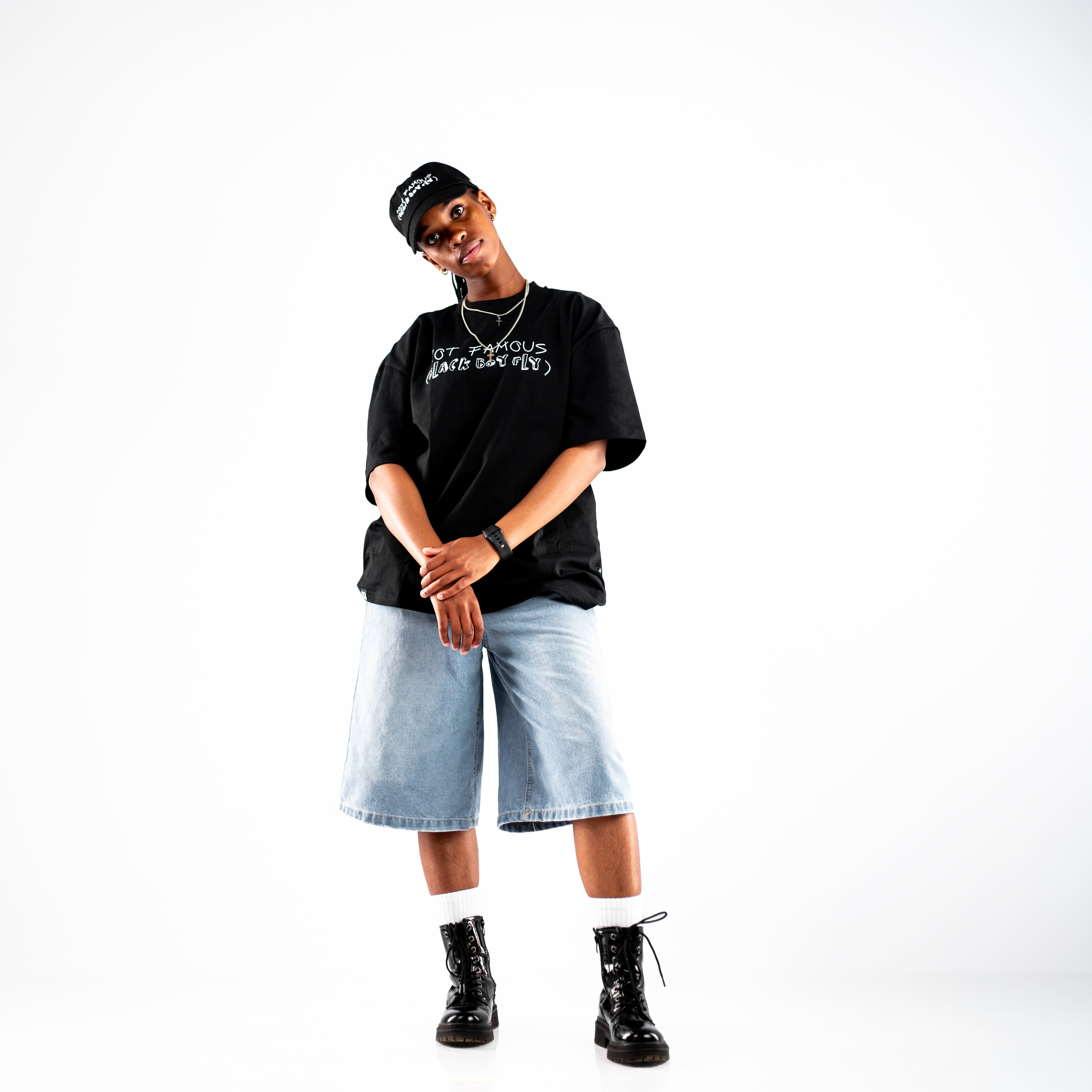 Person standing against a white background wearing a black cap, Kane Keid black t-shirt with white text "Not Famous (Black Boy Fly)", light blue denim shorts, black boots with white socks, and accessories including a watch and necklace. Casual pose with hands clasped in front.