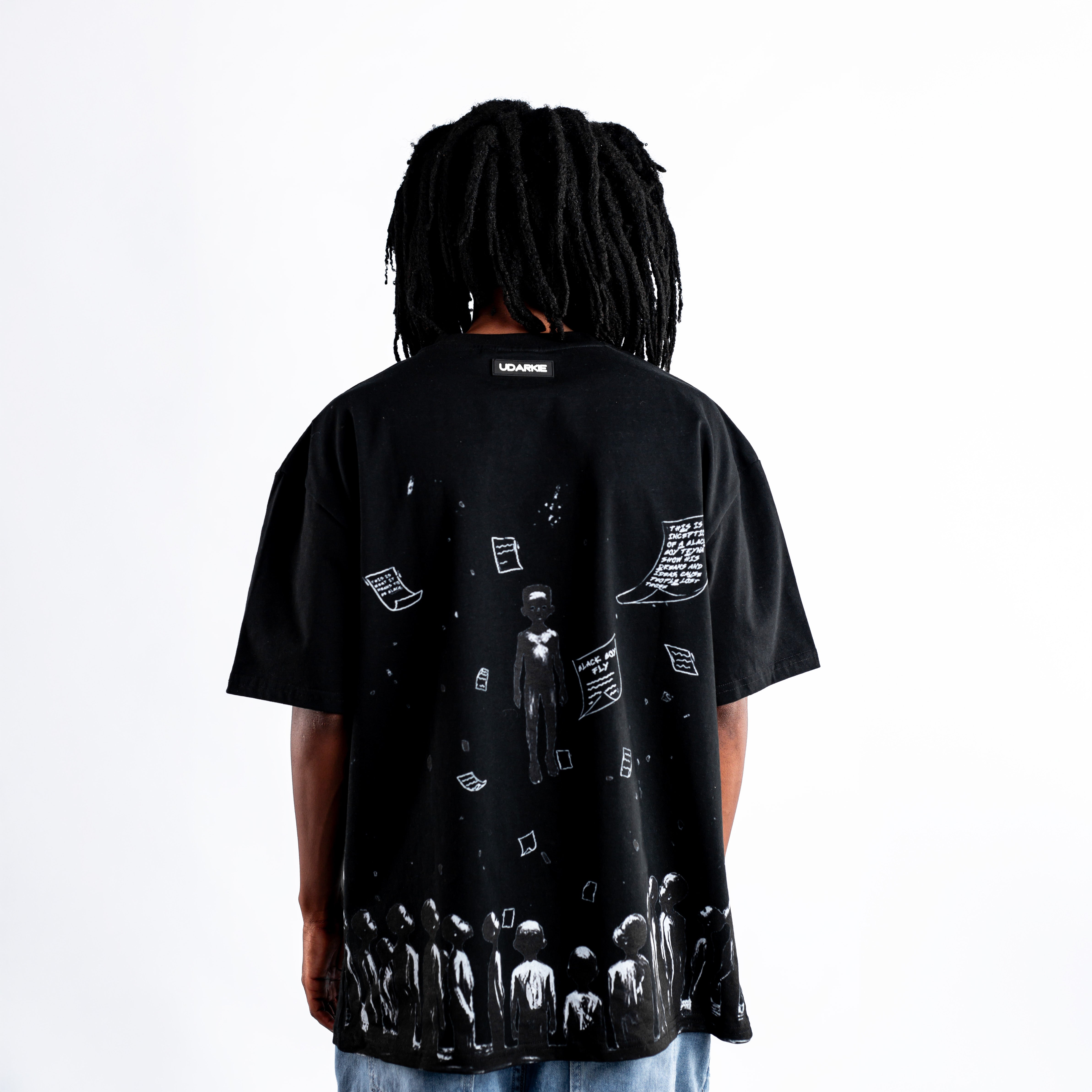 Back view of a person wearing a black Kane Keid Not Famous Black Boy Fly T-shirt featuring an artistic design with figures looking upward, floating sheets of paper, and a central elevated figure. The design is detailed and visually striking.