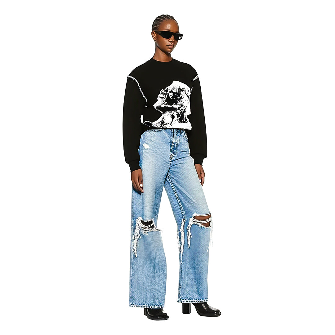 Person wearing a black long-sleeve shirt with white graphic design, light blue distressed wide-legged jeans with large rips, black heeled boots, and dark sunglasses. Set against a plain white background.