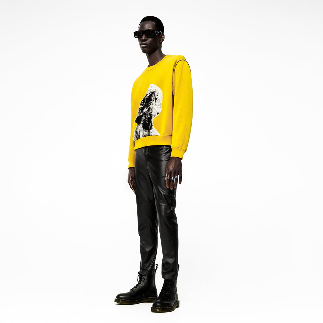Model wearing yellow graphic sweatshirt, black leather pants, black boots, and sunglasses against white background