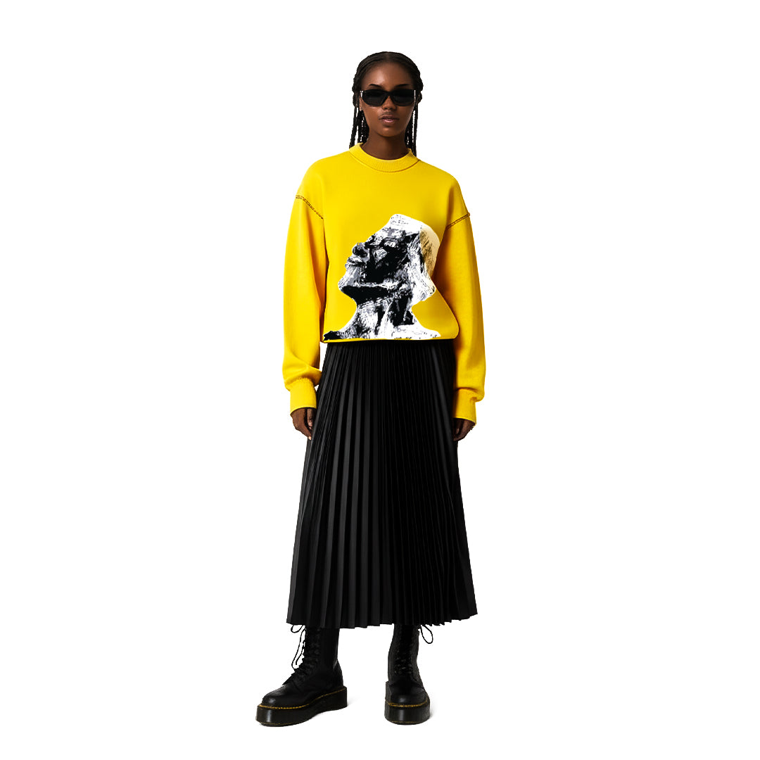 Model wearing yellow graphic sweatshirt with face print, black pleated skirt, black boots, and sunglasses