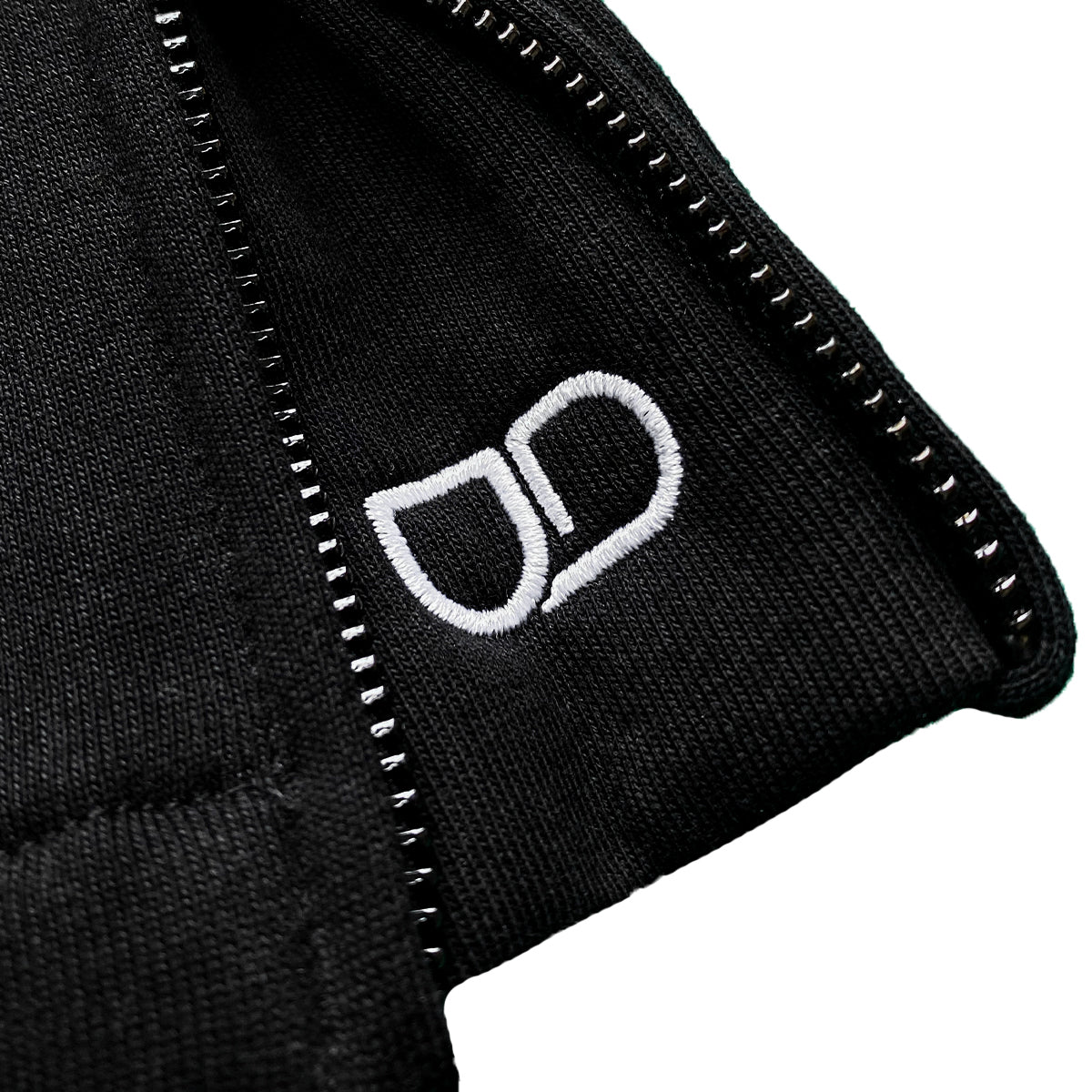 Close-up of black trackpant with a zipper at the bottom and white embroidered "UD" logo near the edge.
