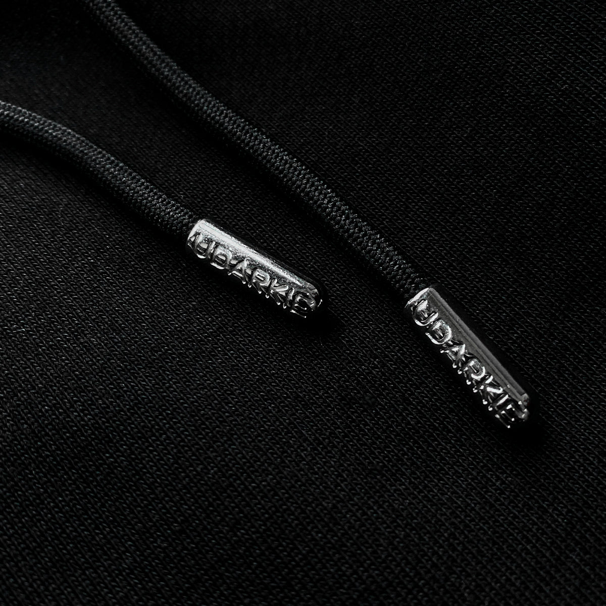 Close-up of black drawstrings with metallic aglets engraved with "UDARKIE" on black fabric
