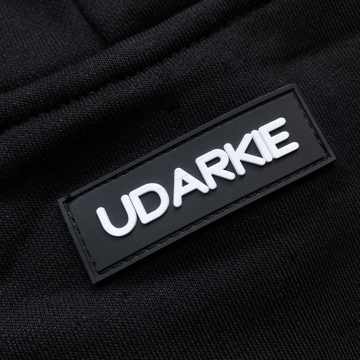 Close-up of black hoodie with rectangular black patch sewn on, displaying "UDARKIE" in raised white letters