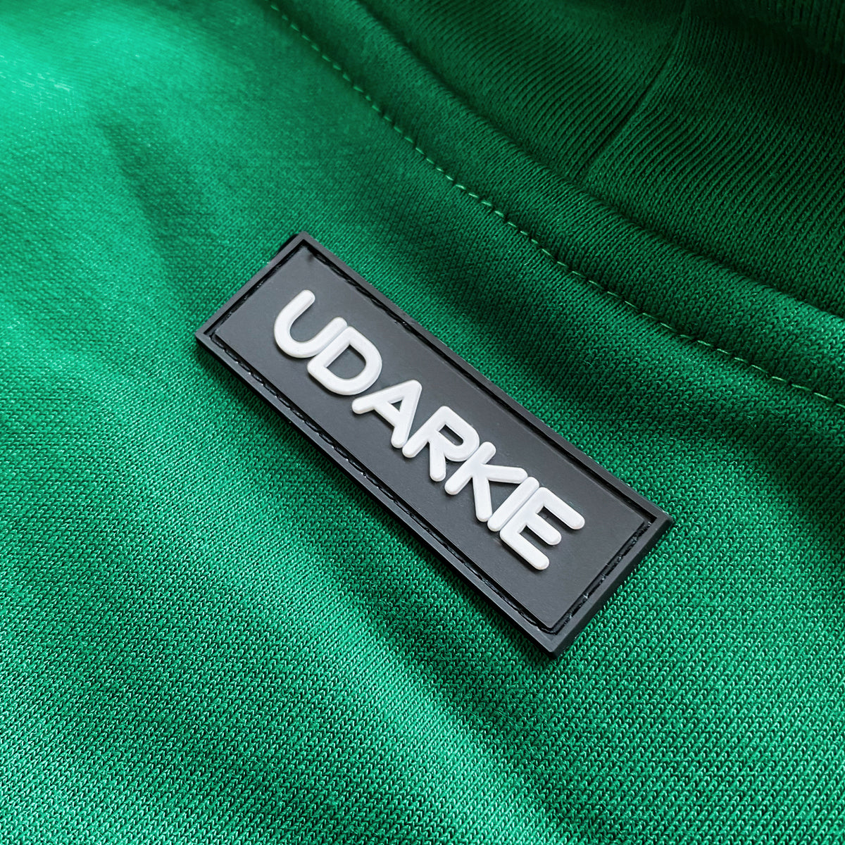 Close-up of green hoodie with rectangular black patch sewn on, displaying "UDARKIE" in raised white letters