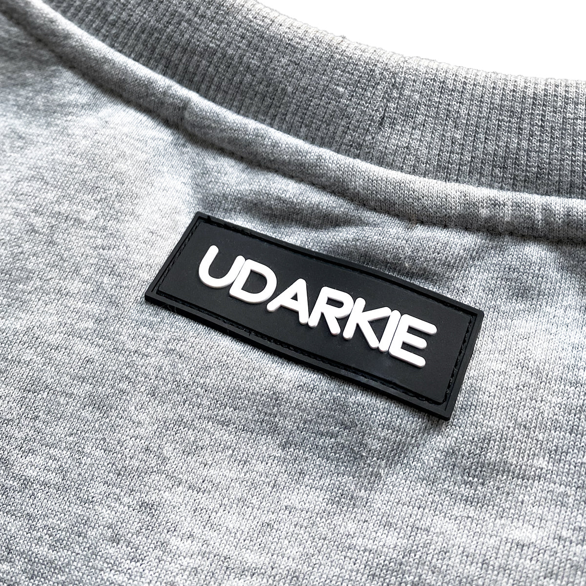 Close-up of gray sweatshirt collar area with black rectangular patch sewn onto the fabric, displaying "UDARKIE" in raised white letters