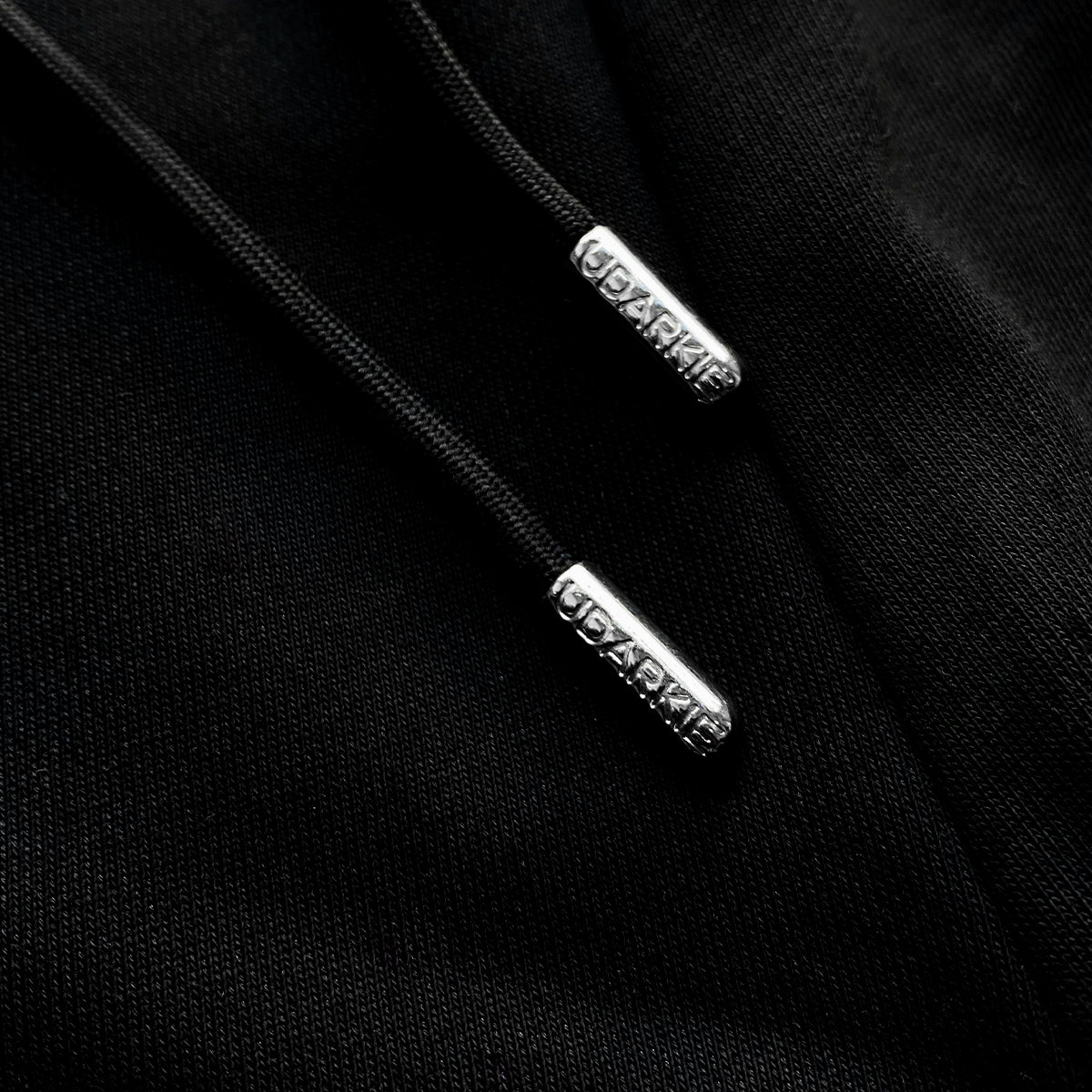 Close-up of black drawstrings with metallic aglets engraved with "UDARKIE" on black fabric, likely part of a sweatpant.