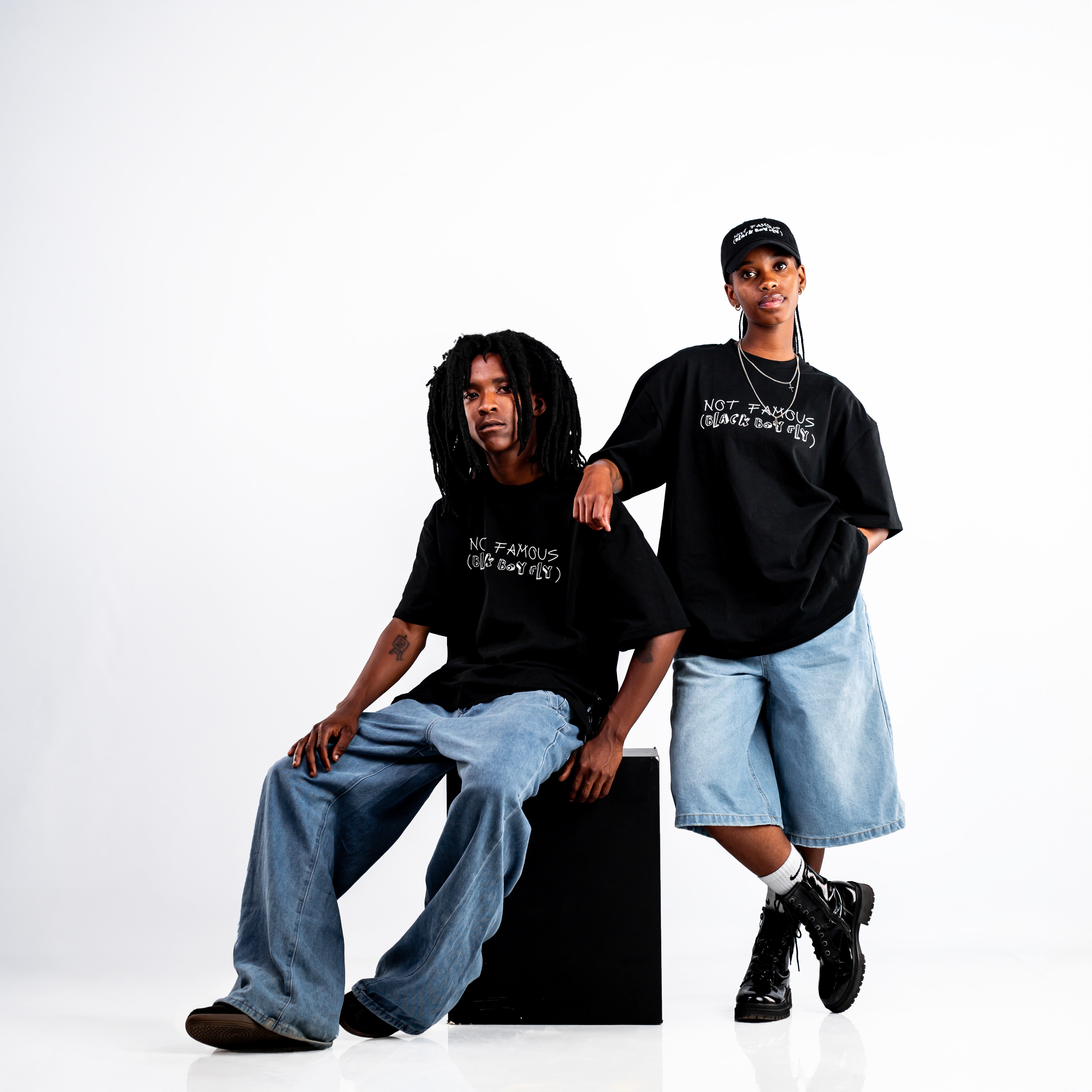 Two individuals showcasing trendy streetwear outfits, featuring Kane Keid Not Famous Black Boy Fly black graphic t-shirts and denim jeans. One person is seated on a black box wearing wide-leg jeans, while the other stands with hands in pockets, wearing knee-length denim shorts and a black cap.