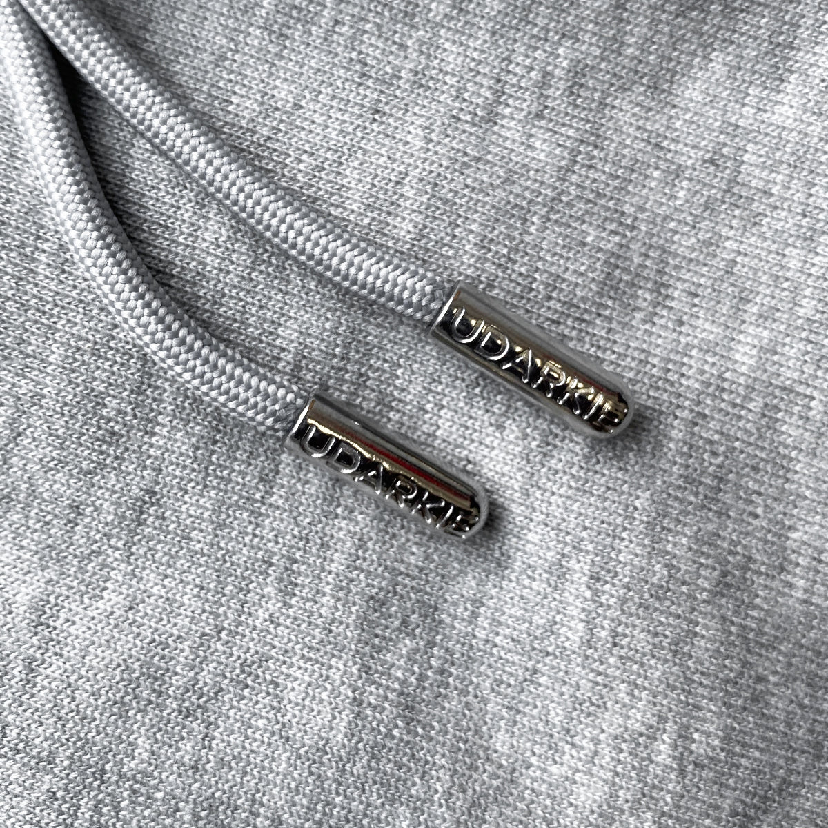 Close-up of gray drawstrings with metallic aglets engraved with "UDARKIE" on black fabric