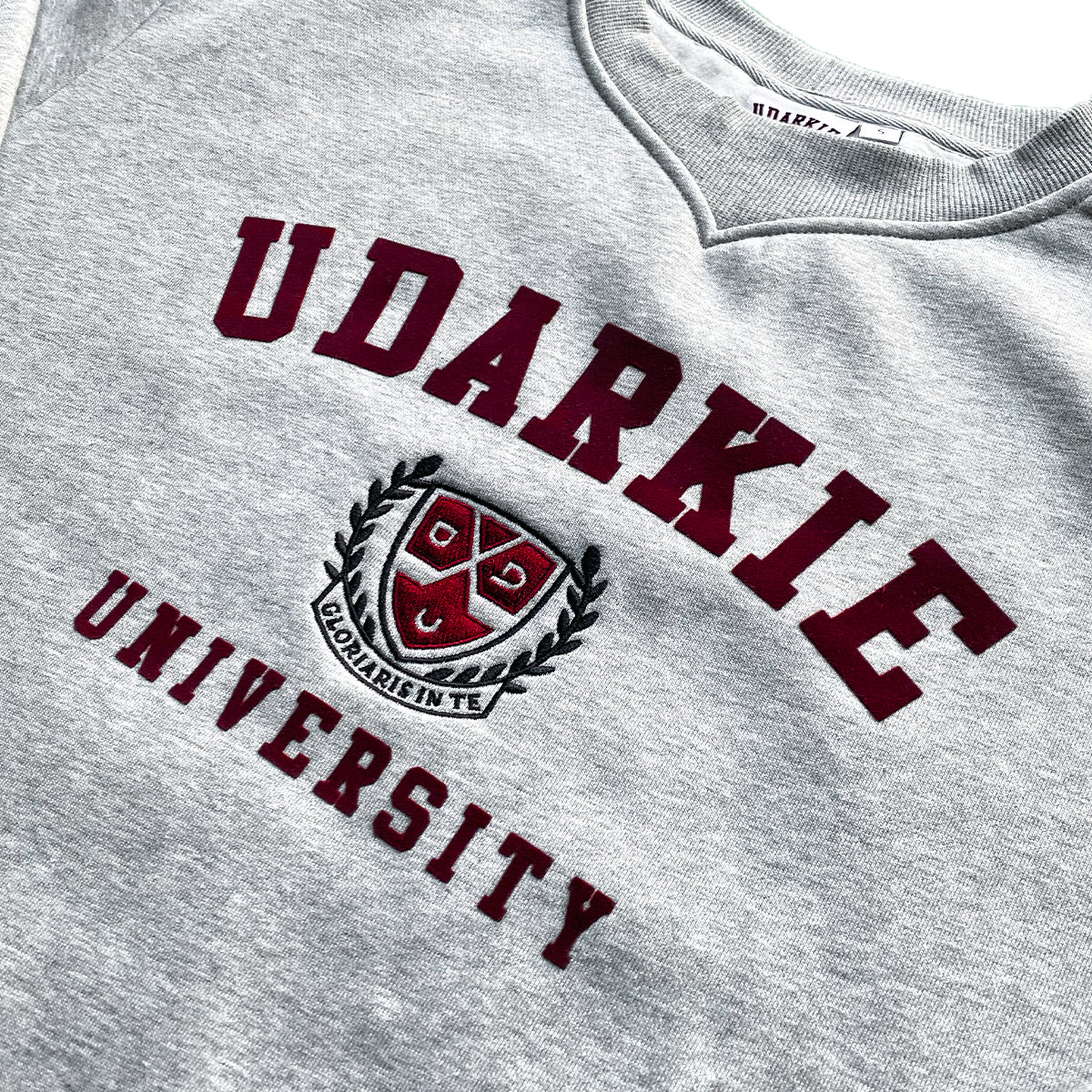 Flat lay of gray sweatshirt with "UDARKIE UNIVERSITY" in maroon letters and embroided badge.